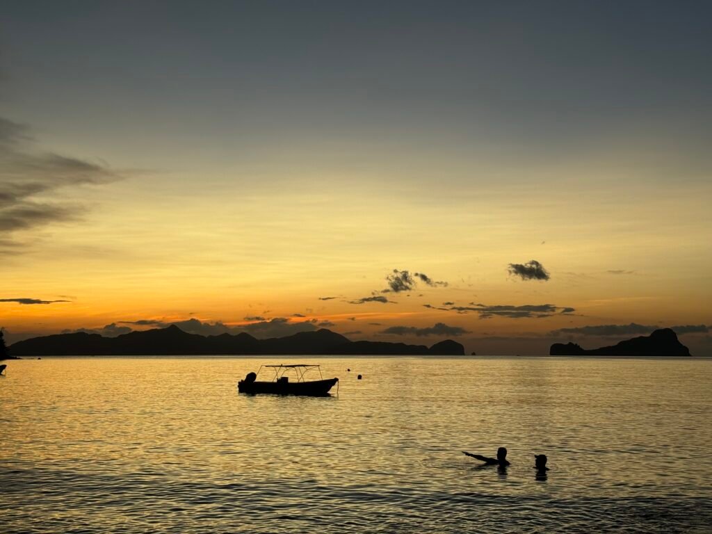 Two men swimming near a boat at Marimegmeg Beach during sunset.